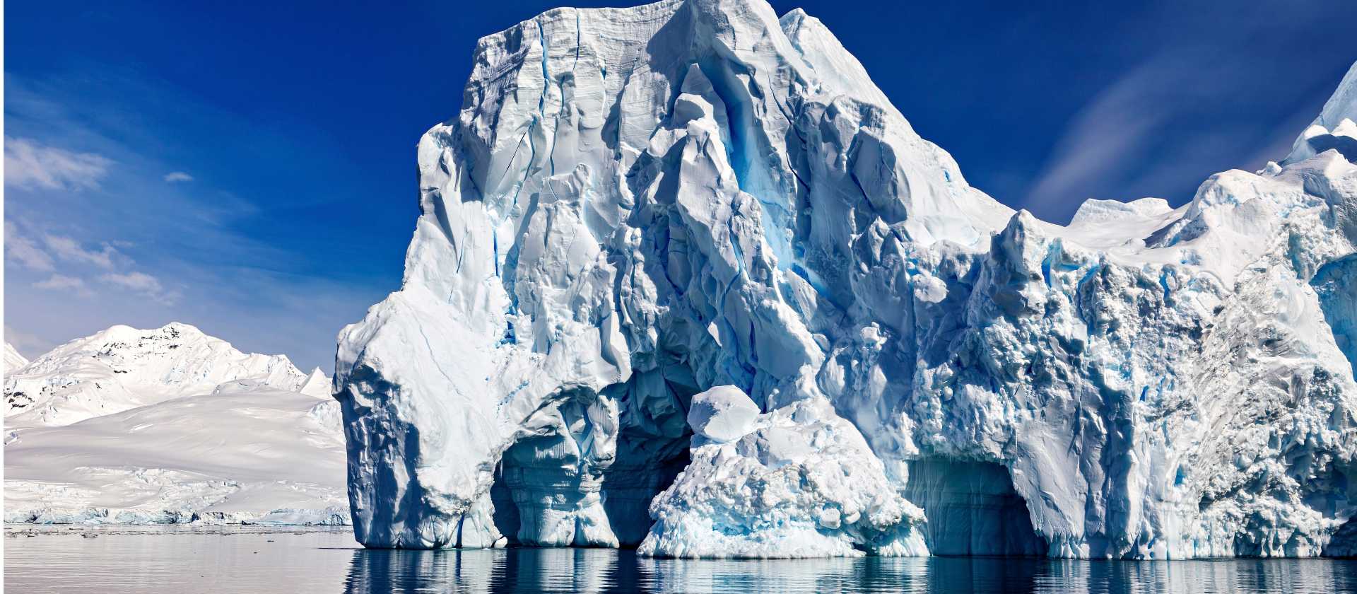Antarctica's Southern Ocean iceberg towers above calm reflective water near snowy peaks.