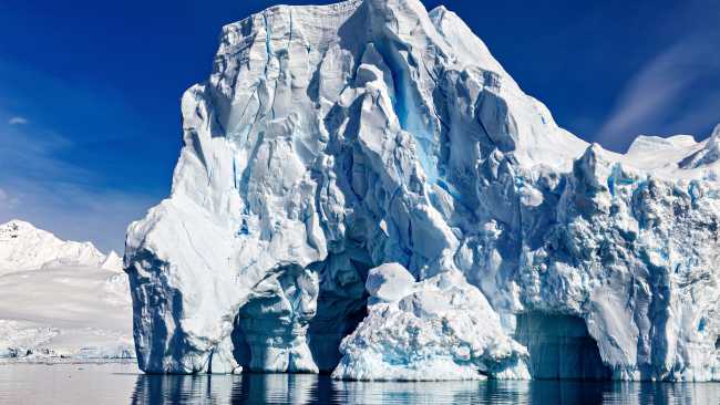 Antarctica's Southern Ocean iceberg towers above calm reflective water near snowy peaks.