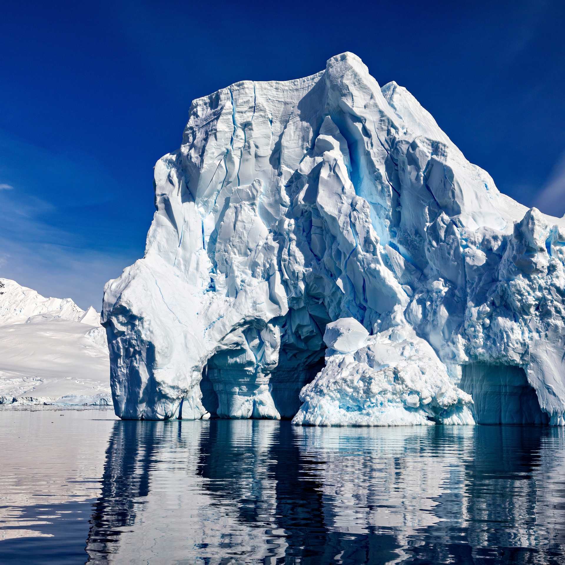 Antarctica's Southern Ocean iceberg towers above calm reflective water near snowy peaks.